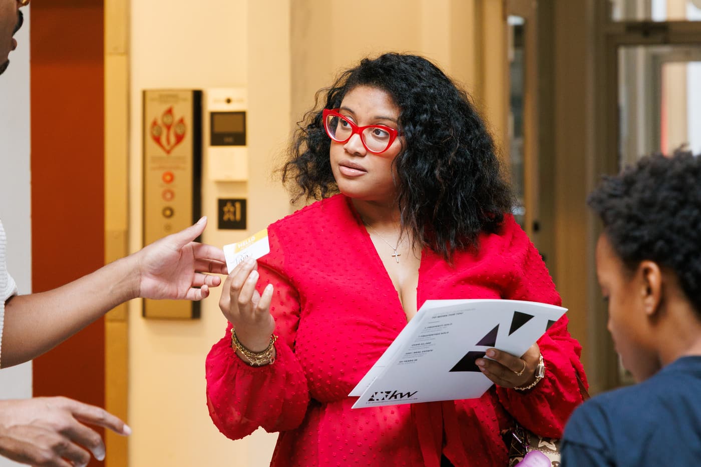 A host in a red dress holding an event program and talking with guests at Stem & Vine, Baltimore